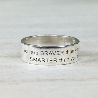 A Sterling Silver band ring with an engraved message on it, displayed against a wooden background.