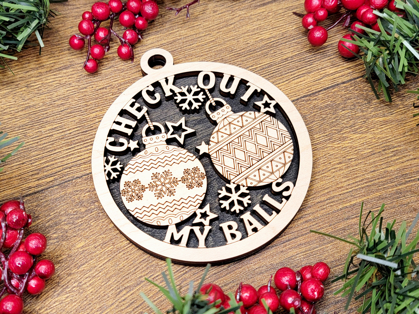 Wooden Christmas ornament with 'Check Out My Balls' text on a wooden surface with berries and greenery.