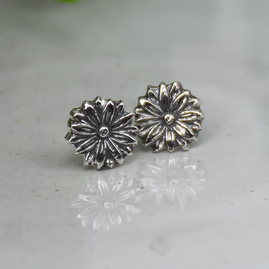 A pair of silver-toned flower-shaped stud earrings with textured petals, resting on a light gray marble surface with a soft green plant leaf blurred in the background.