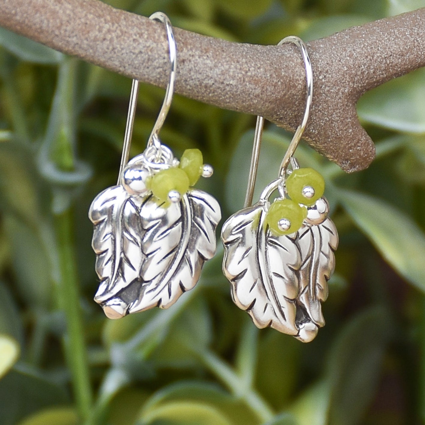 A pair of silver leaf dangle earrings with small lime green bead accents, hanging from a brown branch with a blurred green foliage background.