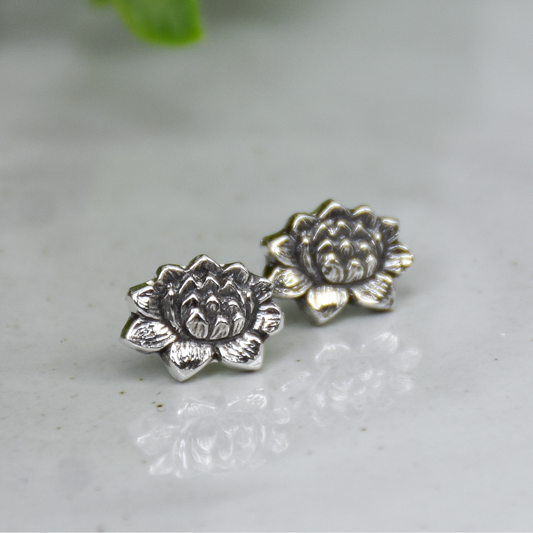 A pair of silver-toned lotus flower stud earrings resting on a light gray marble surface with a soft-focus green plant in the background.