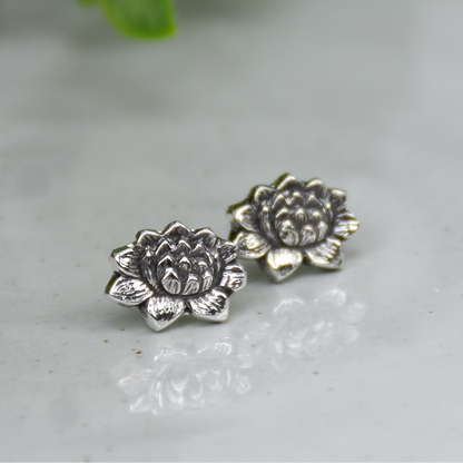 A pair of silver-toned lotus flower stud earrings resting on a light gray marble surface with a soft-focus green plant in the background.